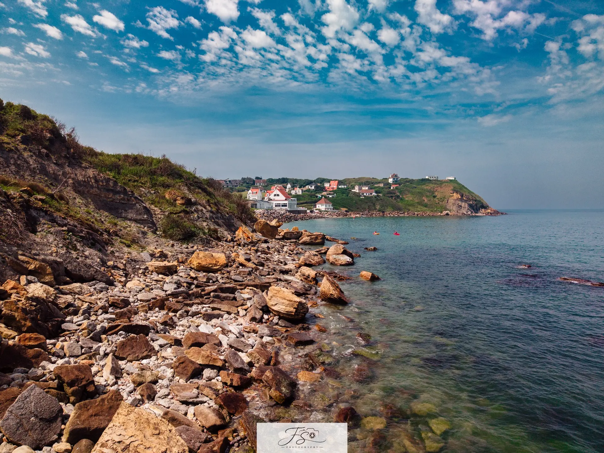 Côte minérale — rivage rocheux et village côtier sous un ciel bleu ponctué de nuages