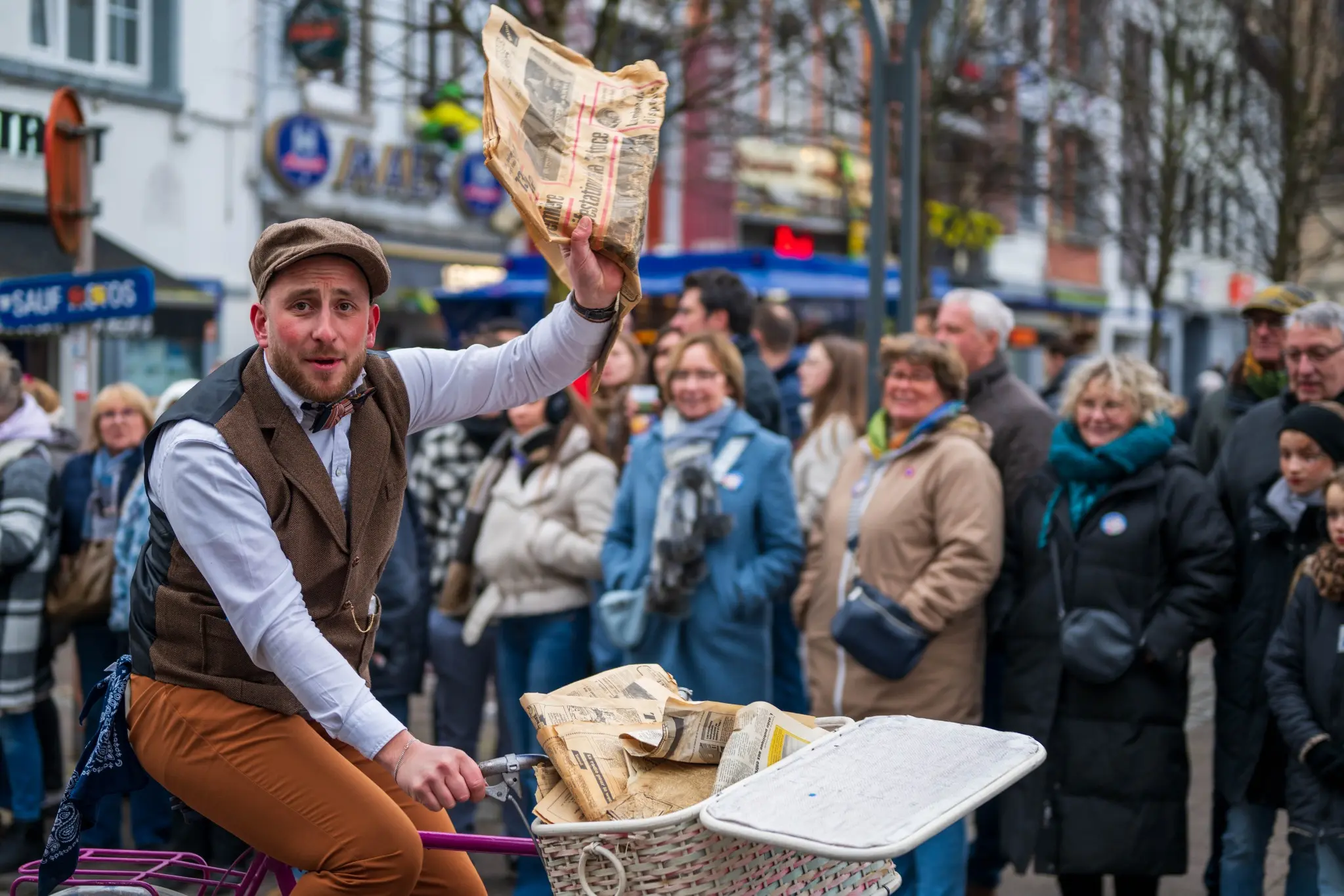 Animateur costumé à vélo brandissant un journal devant le public dans une rue de Malmedy